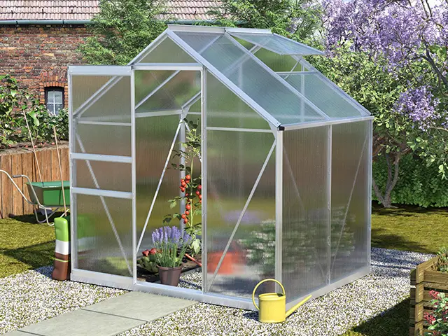 Ein Gewächshaus steht im Garten, beherbergt Tomatenpflanzen und ist von Blumen umgeben.