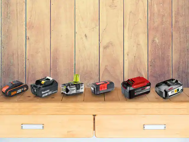 Sechs Akkus stehen nebeneinander auf einem Holztisch vor einer Holzpaneelwand.