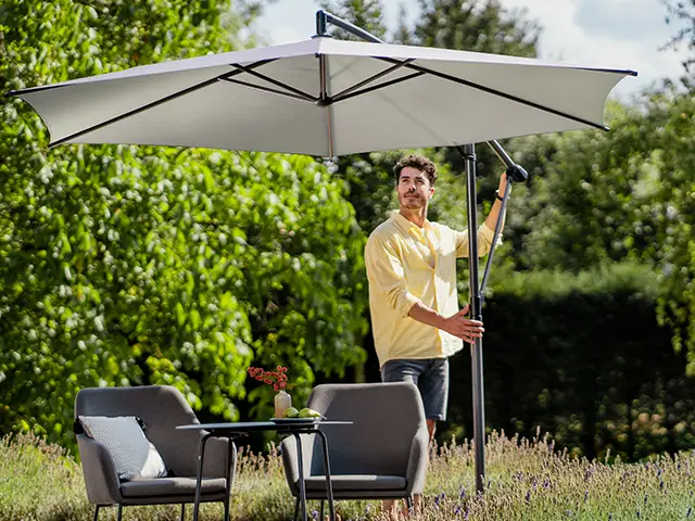 Ein Mann öffnet einen großen Sonnenschirm neben Gartenmöbeln in einem grünen Außenbereich.