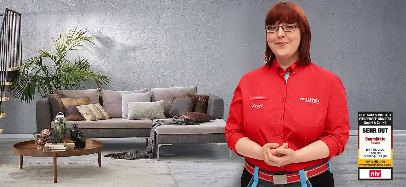 Eine lächelnde Hellweg Mitarbeiterin steht in einem modernen Wohnzimmer mit Sofa und Pflanzen.