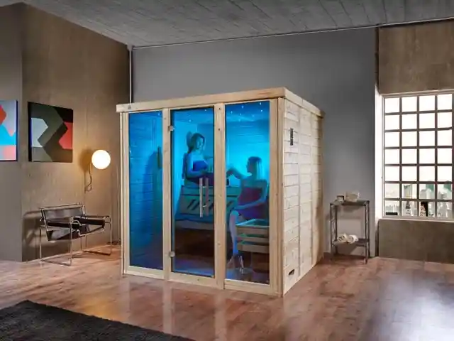 Zwei Personen sitzen in einer beleuchteten Sauna in einem modernen Raum mit Bildern.