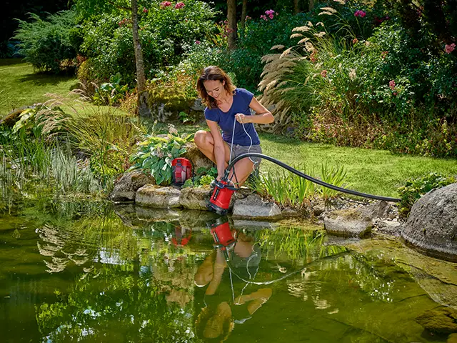 Eine Person reinigt mit einer Einhell Pumpe einen Teich in einem bepflanzten Garten.