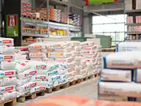 Paletten mit Zementsäcken stehen gestapelt in einem Baumarkt vor Regalen voller Baumaterialien.