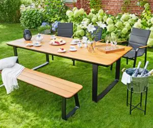 Ein Holztisch steht im Garten mit gedecktem Geschirr, umgeben von Stühlen und Pflanzen.