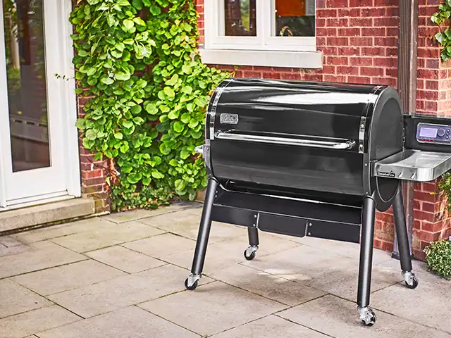 Ein schwarzer Pellet-Smoker steht auf einer Terrasse vor einer roten Backsteinmauer mit Pflanzen.