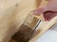Eine Hand streicht mit einem Pinsel Holz in einer Werkstatt.