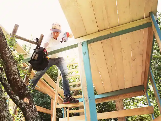 Ein Mann baut ein Baumhaus mit einem Werkzeug inmitten von Bäumen.