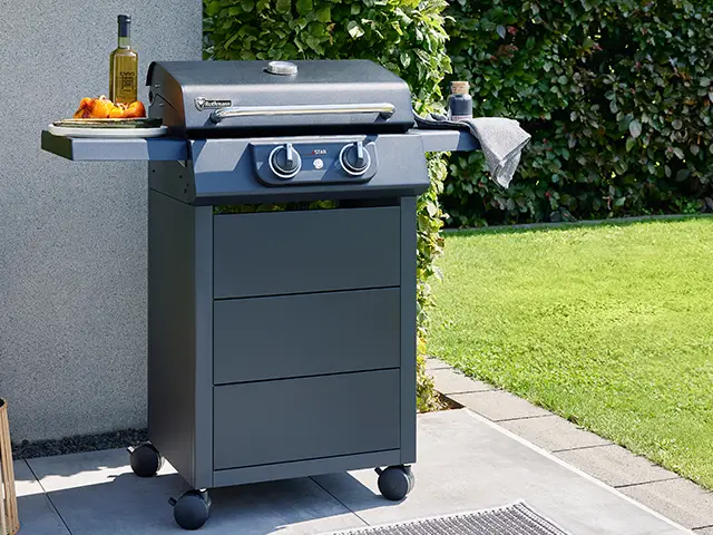 Ein Gasgrill steht draußen auf einer Terrasse neben einer Hecke und grünem Rasen.