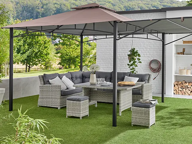 Ein Pavillon mit Gartenmöbeln steht auf einem Rasen vor einem Haus mit weißer Wand und Pflanzen.