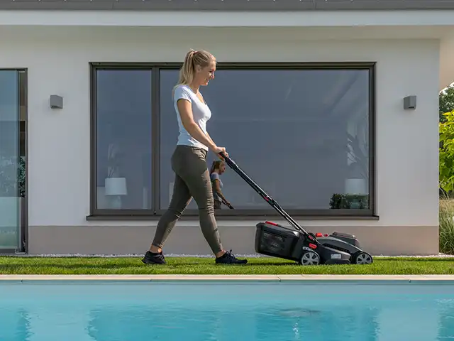 Eine Frau mäht den Rasen neben einem modernen Haus mit großen Fenstern und Pool.