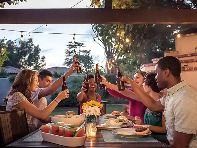 Sechs Personen stoßen an einem gedeckten Tisch mit Bierflaschen im Garten an.