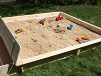 Ein großer Sandkasten mit Spielzeug steht auf einer Rasenfläche im Freien.