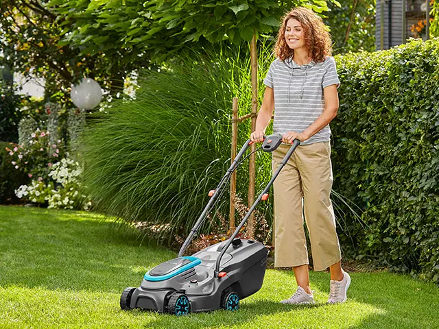 Eine Person schiebt einen Rasenmäher in einem grünen Garten mit Büschen und Bäumen.