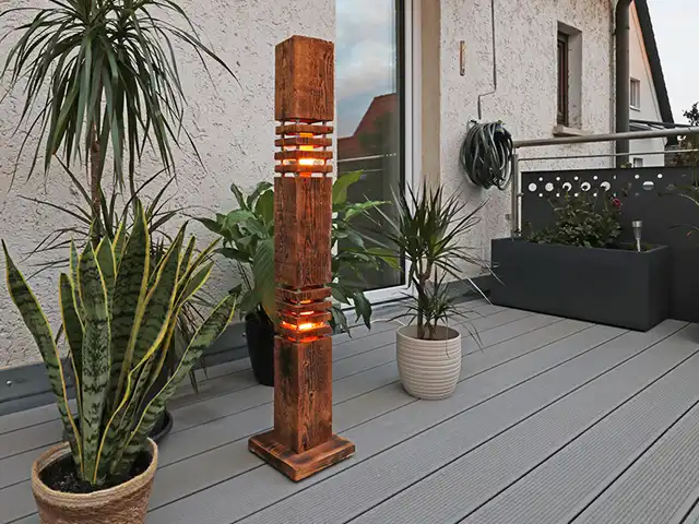 Eine Holzlampe leuchtet auf einem Balkon neben Pflanzen in Töpfen und einem grauen Dielenboden.