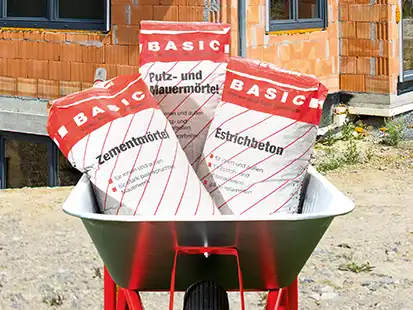 Eine Schubkarre mit drei Zementsäcken steht vor einem im Bau befindlichen Ziegelhaus.