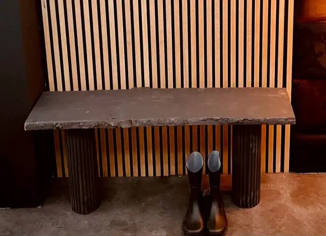 Eine schwarze Sitzbank steht vor einer Holzwand, daneben stehen Gummistiefel auf dem Boden.