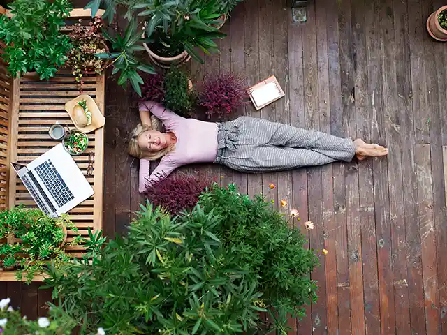 Eine Frau liegt entspannt auf einem Holzboden, umgeben von Pflanzen und einem Laptop auf einer Bank.