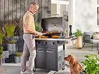 Ein Mann grillt Fleisch auf einem Grill im Garten; ein Hund sitzt daneben.