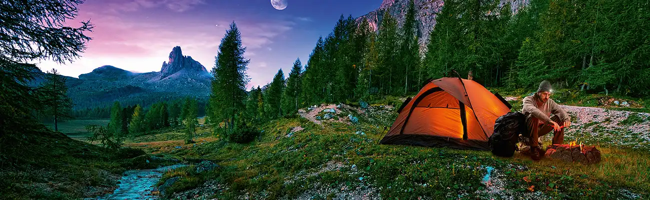 Ein oranges Zelt leuchtet im Wald neben einem Lagerfeuer, umgeben von Bergen und Bäumen.
