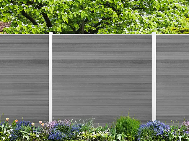 Ein grauer Zaun steht vor blühenden Blumen, mit grünen Bäumen im Hintergrund.