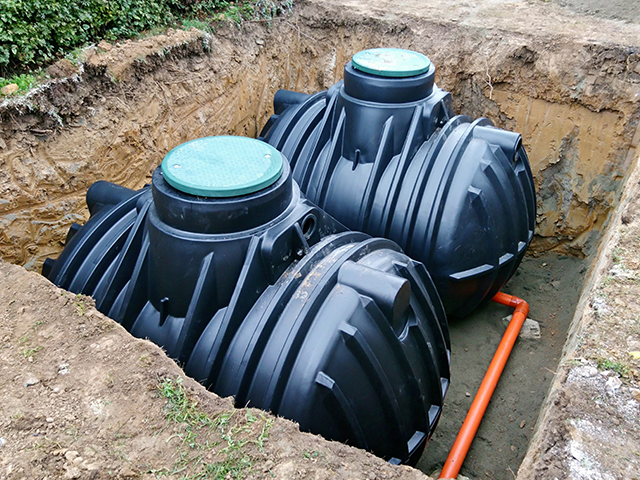 Zwei schwarze Wassertanks sind in einem Erdloch vergraben und mit Rohrleitungen verbunden.