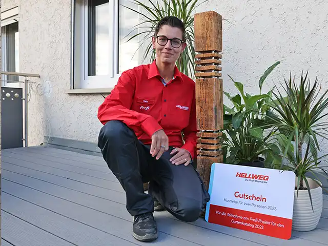 Eine Mitarbeiterin kniet neben einer Holzskulptur auf einer Terrasse. Neben ihr ein HELLWEG Gutschein für einen Kurzurlaub.