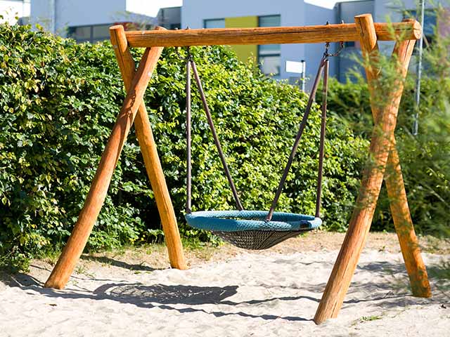 Eine Nestschaukel hängt ruhig in einem sonnigen Spielplatz, umgeben von grünen Büschen.