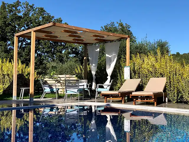 Zwei Liegestühle stehen neben einer Pergola hinter einem Pool in einem sonnigen Garten.