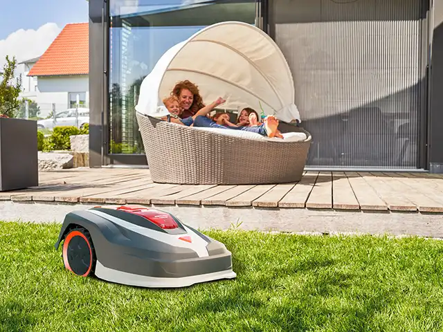 Ein Roboter-Rasenmäher mäht den Rasen, während zwei Personen in einer Gartenliege entspannen.