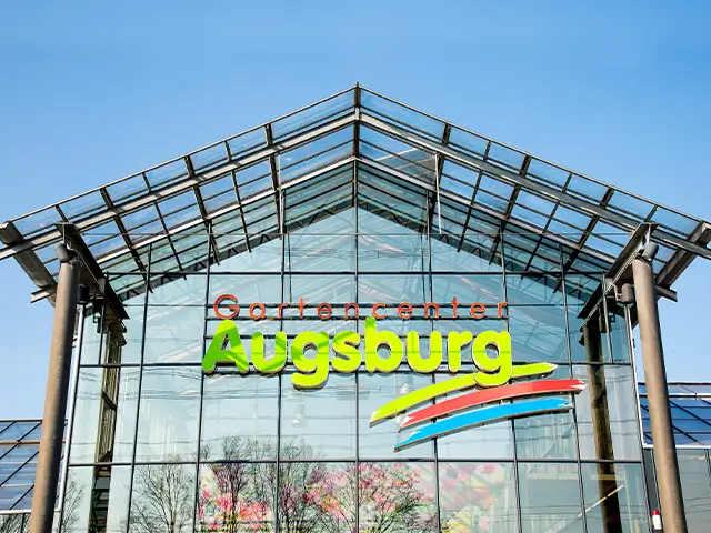 Gewächshaus mit Schriftzug "Gartencenter Augsburg" steht unter blauem Himmel.