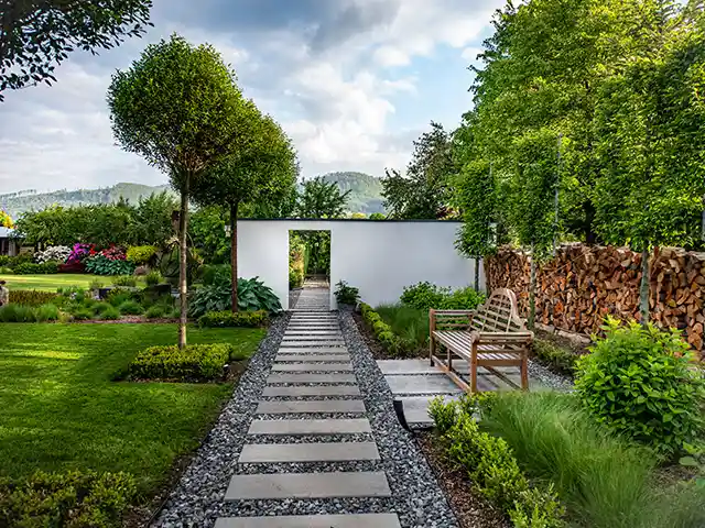 Ein gepflasterter Weg führt durch einen grünen Garten mit einer Bank und einer Holzwand.