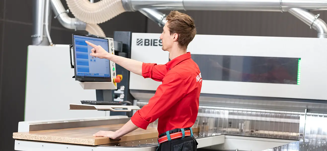 Ein Mann in einem roten Hemd bedient einen Computerbildschirm in einer industriellen Werkstattumgebung.