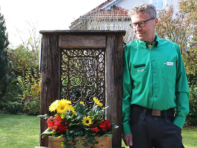 Ein BayWa-Mitarbeiter lehnt neben einem Holzgitter mit Blumen in einem grünen Garten