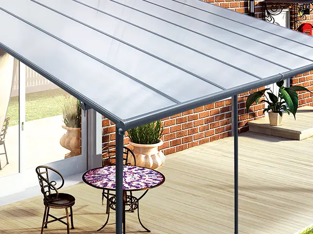 Ein Tisch und Stühle stehen unter einem Vordach auf einer Holzterrasse neben einem Backsteinhaus.