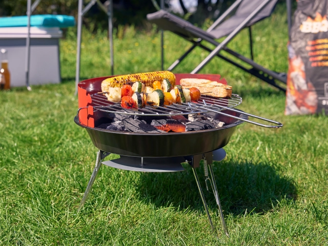 Holzkphlegrill mit Genüsespießen auf einer Wiese beim Campen