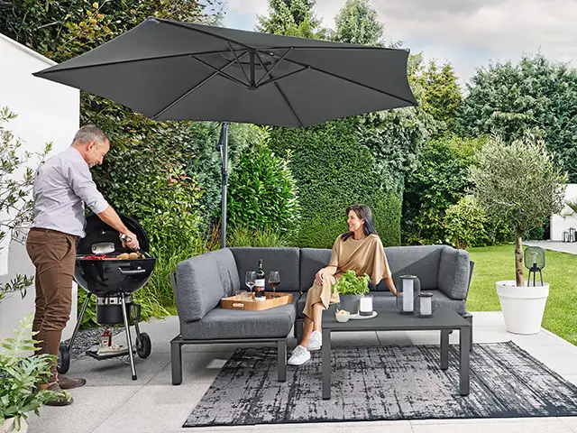 Ein Mann grillt, während eine Frau auf einem grauen Sofa im Garten sitzt.