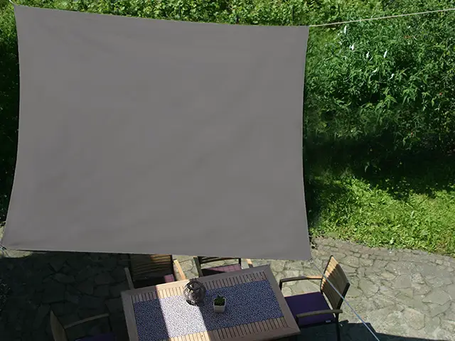 Ein graues Sonnensegel hängt über einem Tisch mit Stühlen im Garten bei sonnigem Wetter.