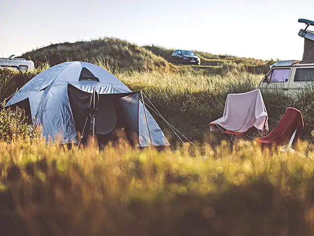 Ein Zelt steht neben Campingstühlen in einer grünen Wiese mit Autos im Hintergrund.