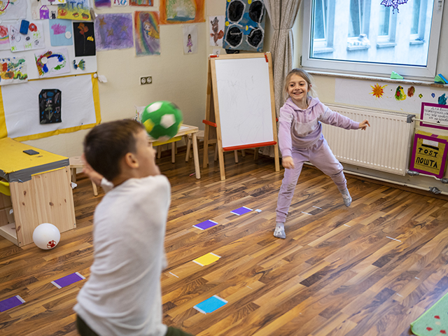 Zwei Kinder spielen mit einem Ball in einem lebhaft dekorierten Klassenraum.