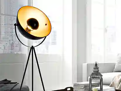 Eine Stehlampe leuchtet in einem modern eingerichteten Wohnzimmer neben einer Couch und einem Bücherstapel.