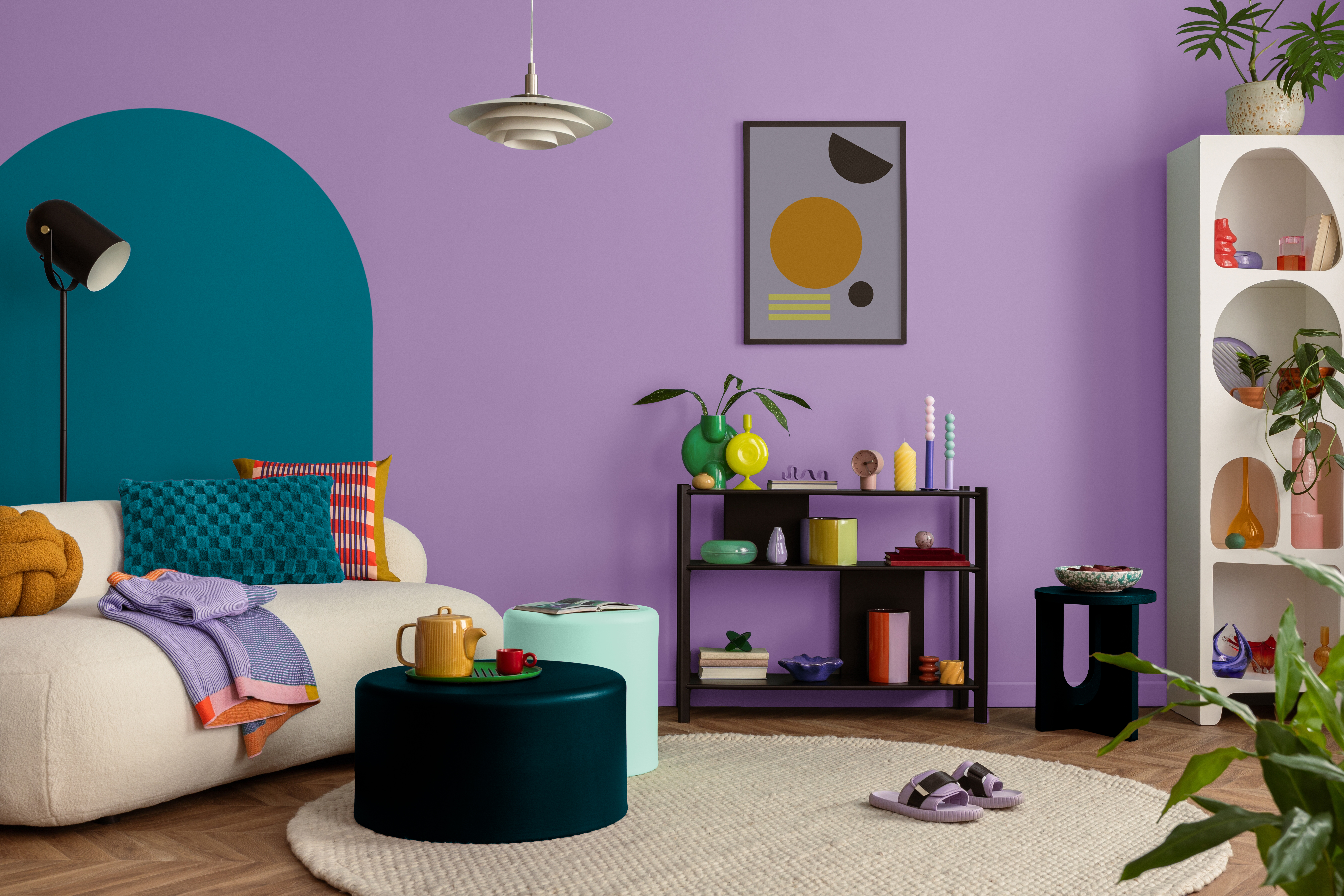 Helles Sofa vor einer lila Wand. Lila Schuhe stehen auf einem beigen Teppich auf einem Holzboden. Die Regale sind weiß und schwarz.