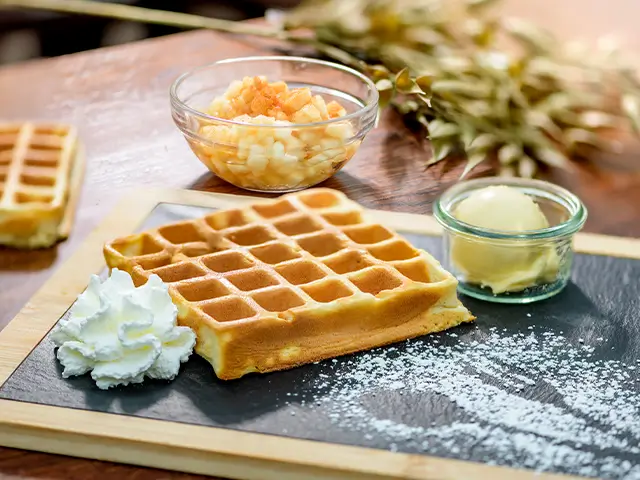 Eine frische Waffel liegt neben Sahne, Eis und Apfelstücken auf einem Tisch.