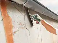 Eine Hand verputzt eine Wand mit einer Kelle unter einer Regenrinne im Freien.
