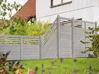 Ein grauer Zaun steht in einem Garten vor einem weißen Haus mit Pflanzen umgeben.