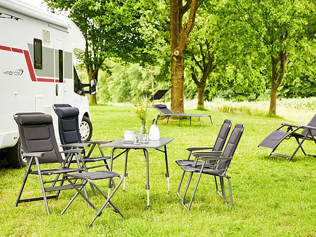 Ein Tisch mit Stühlen steht neben einem Wohnmobil auf einer grünen Wiese mit Bäumen.